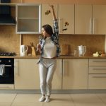 Cheerful young funny woman dancing and singing with ladle while having leisure time in the kitchen at home in the morining