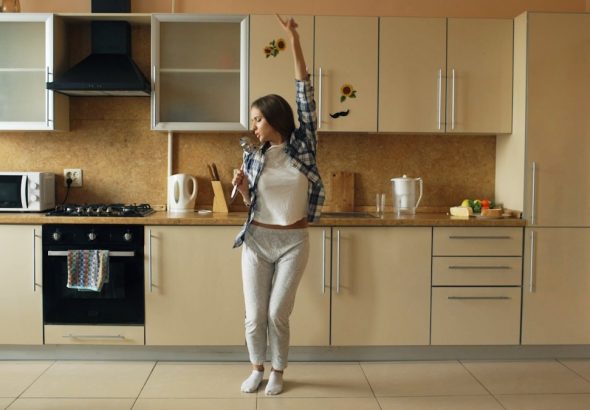 Cheerful young funny woman dancing and singing with ladle while having leisure time in the kitchen at home in the morining