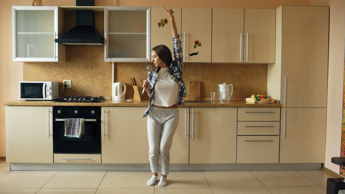 Cheerful young funny woman dancing and singing with ladle while having leisure time in the kitchen at home in the morining
